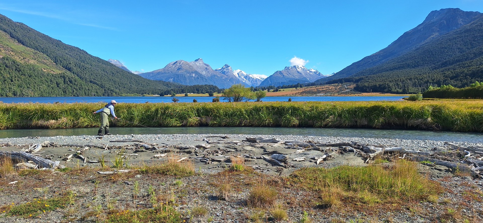 A fly angler casting along a clear backcountry stream with a turquoise lake and snow-capped peaks behind